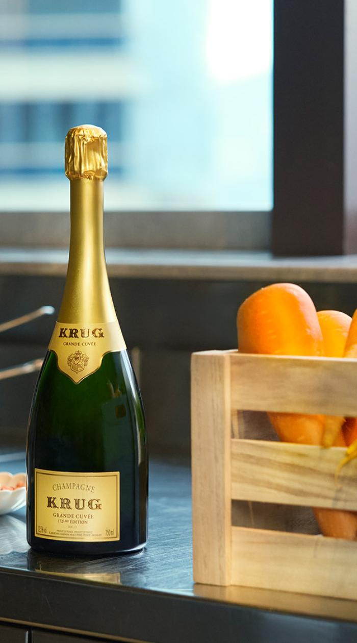 A bottle of Krug champagne next to a wooden crate packed with carrots